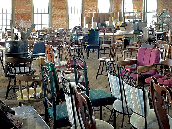 This photo is taken from one side of an old factory room with brick walls and wooden floors, bright white light glows in through tall industrial windows. The room is filled with messy rows of dining chairs of many varietes and along the wall across the room there are stacks of lampshades, a table, and some shelves. The chairs that fill the space are mostly wooden, some have upholstered seats.